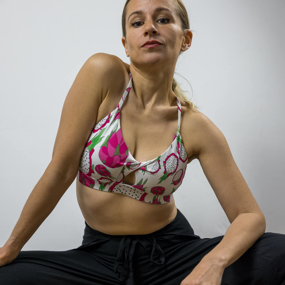 Woman squatting wearing a white and pink sports bra with a twisted cutout detail at the front. The designer print is a Dragon Fruit or Pitaya fruit. She is wearing black, drawstring pants.